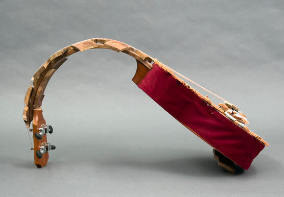 Side view. A ukelele shaped sculpture with a "c" curved neck made of jagged wood material. A ukelele body with red velvet sides and broken ceramic pieces that are white, gold, and maroon glues on the front. Ukelele is strung with copper wire.