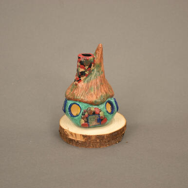 Small cottage made of clay. Brown and green roof, cyan body with dark blue details, red/grey/black stones on door and chimney.