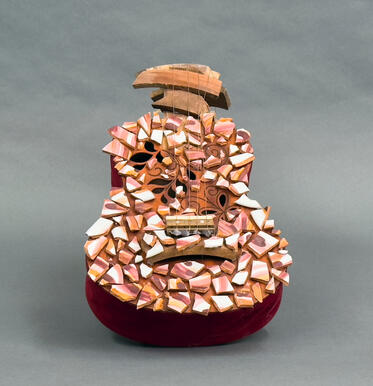Front view. A ukelele shaped sculpture with a "c" curved neck made of jagged wood material. A ukelele body with red velvet sides and broken ceramic pieces that are white, gold, and maroon glues on the front. Ukelele is strung with copper wire.
