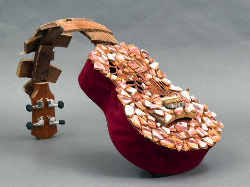 A ukelele shaped sculpture with a "c" curved neck made of jagged wood material. A ukelele body with red velvet sides and broken ceramic pieces that are white, gold, and maroon glues on the front. Ukelele is strung with copper wire.